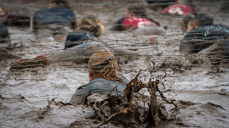 Group of women and men crawl under barbed wire in the mud during a training drill or competition.