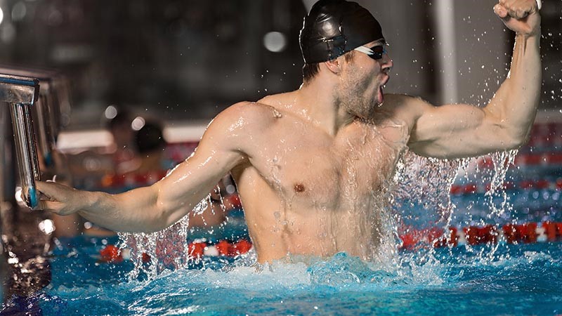 Male swimmer holding block at the edge of a pool and celebrating a win.